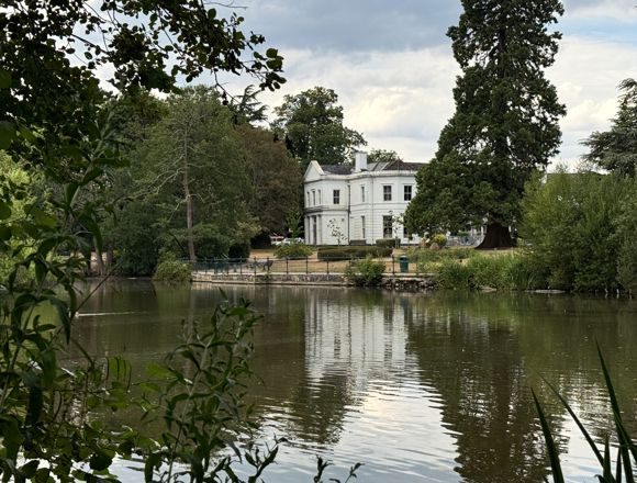 A lake surrounded by trees and greenery with a large white building in the distance.